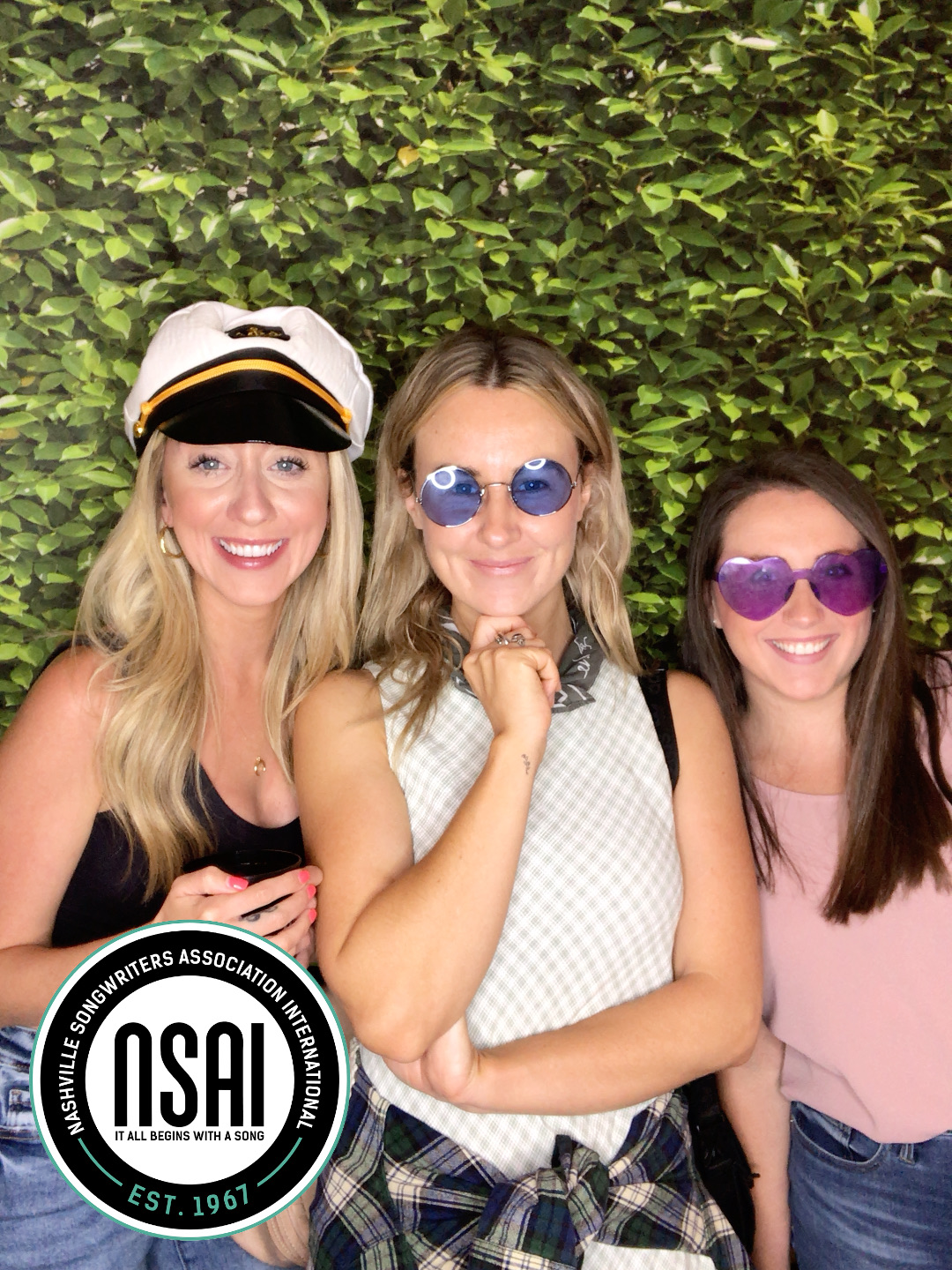 Three smiling ladies posing for a Novabooths photo in front of a greenery backdrop
